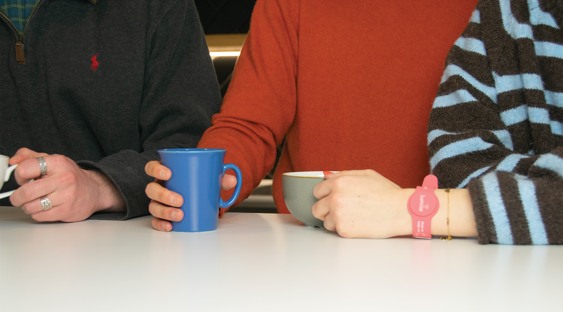 Three people sat at a table with coffee cups, one person has a pink Lumiio safeguarding wristband